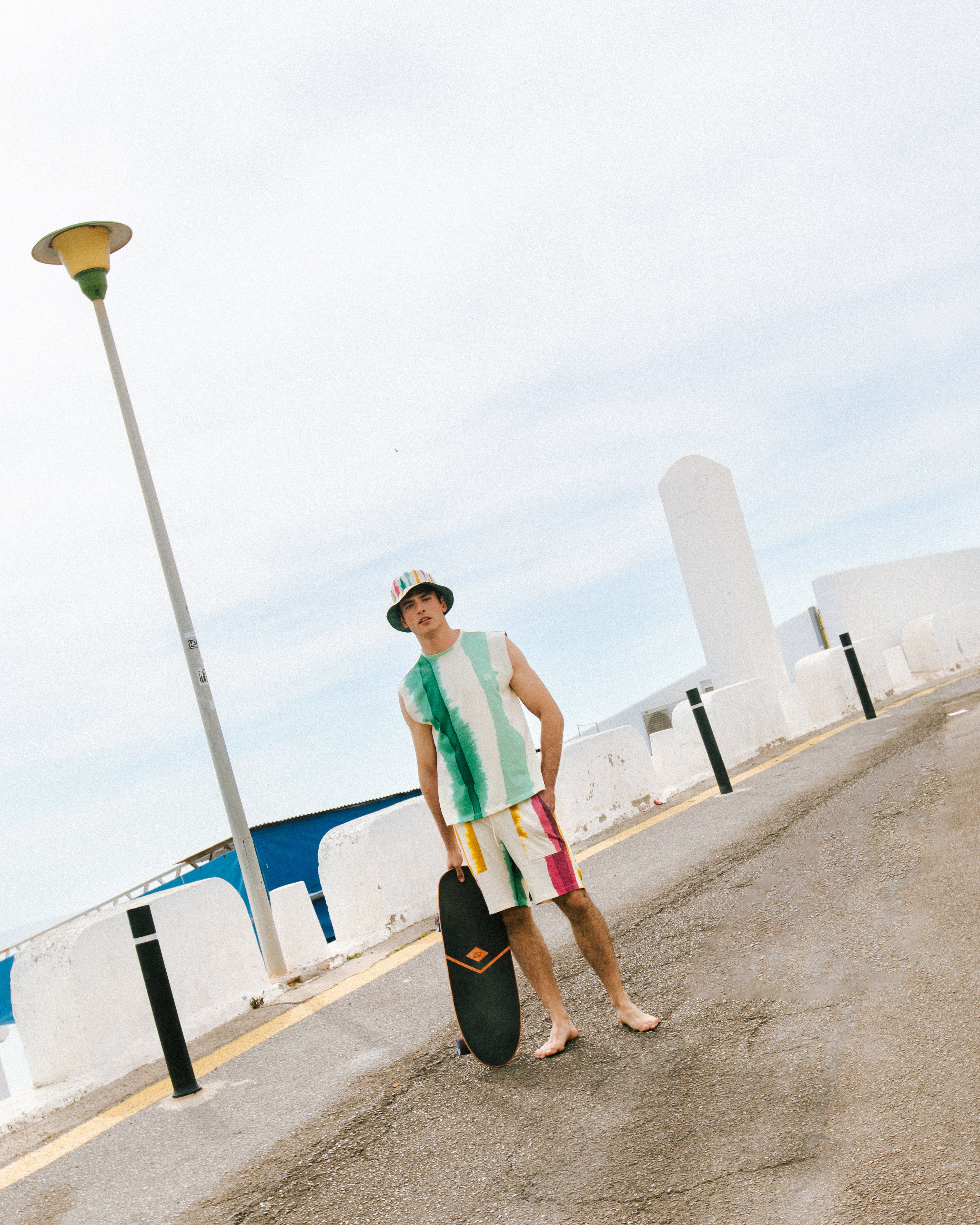 Person holding a skateboard on a road with a white wall and light pole in the background