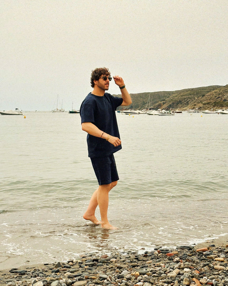 Man standing in Navy outfit on a rocky beach with water and boats in the background