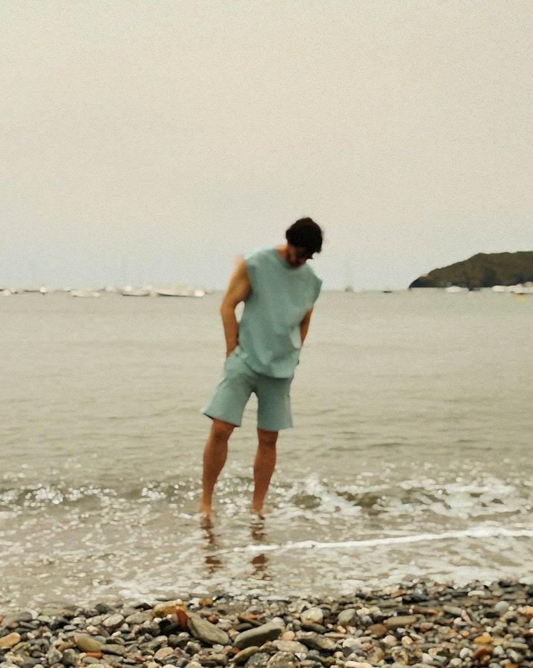 Person in light green outfit standing on a beach with water and rocks in the foreground