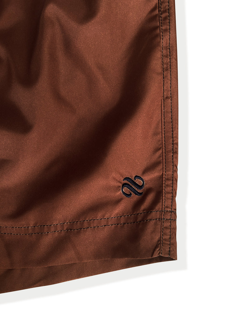 Close-up of a brown fabric garment corner with black embroidered logo and visible stitching on a white background.