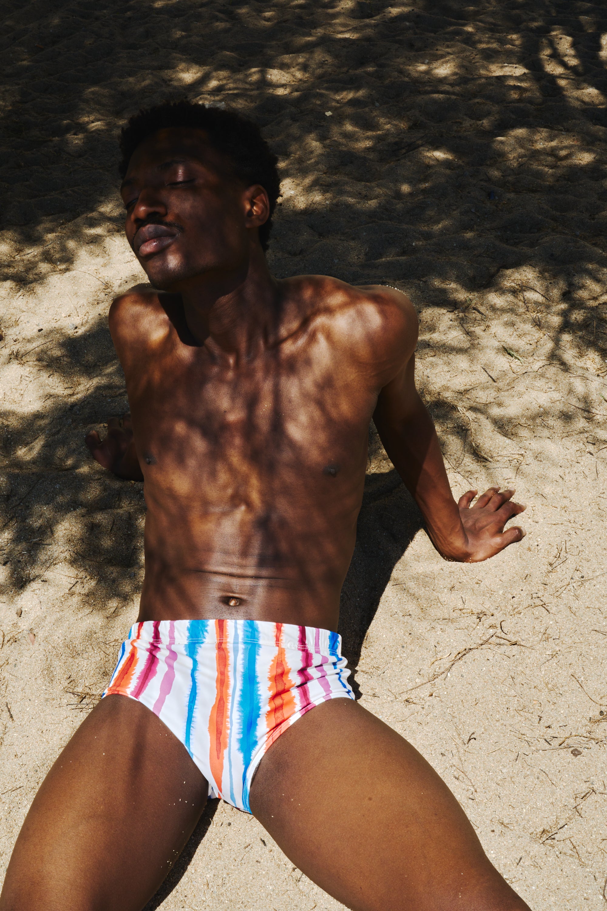 A man lying on sandy ground under dappled tree shadows, wearing low-rise swim briefs with vertical pink, blue, orange, and white stripes.