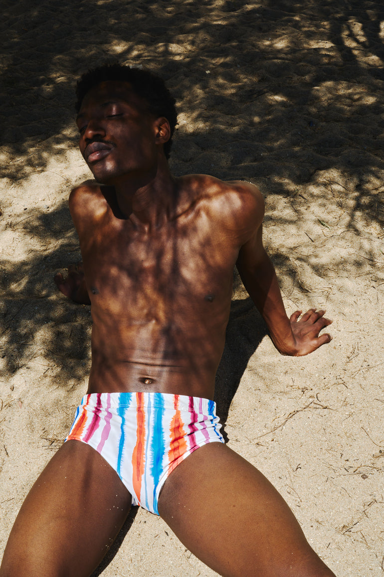 A man lying on sandy ground under dappled tree shadows, wearing low-rise swim briefs with vertical pink, blue, orange, and white stripes.