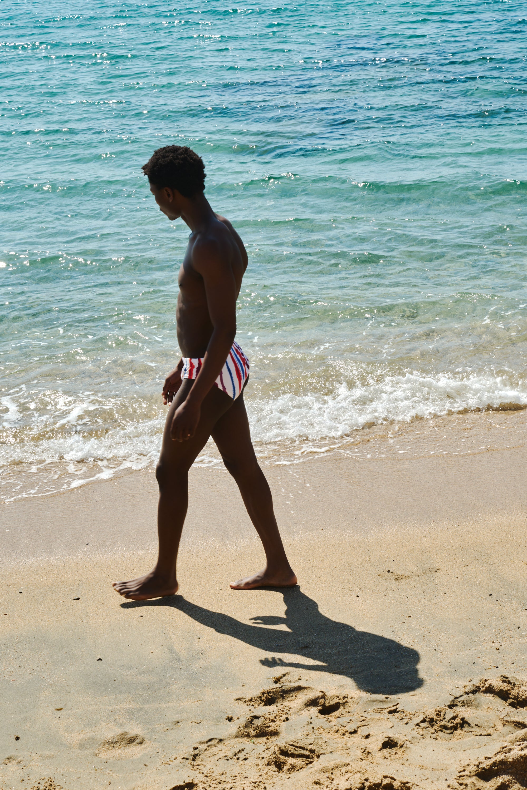 A man walks along a sandy beach next to the ocean, wearing red, white, and blue striped swim briefs.