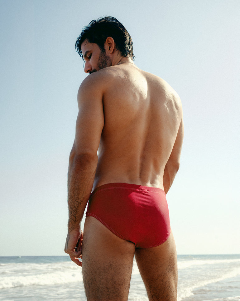 A man standing on a beach viewed from behind, wearing bright red swim briefs against a pale sky and ocean background.