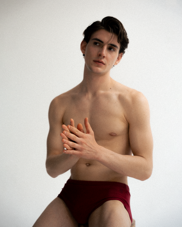 A shirtless man wearing burgundy swim briefs poses against a plain light gray studio background.